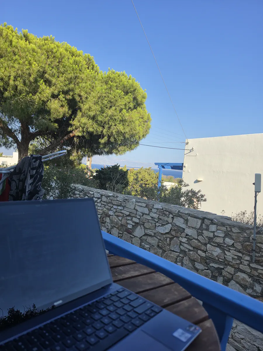 An open laptop on a wooden table on a terrace, with a dry stone wall, a pine tree and the sea beyond