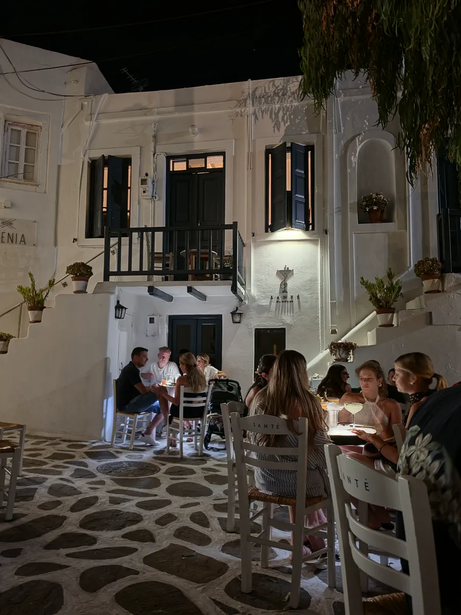 Diners at outdoor tables on a cobbled street between whitewashed Cycladic houses at night